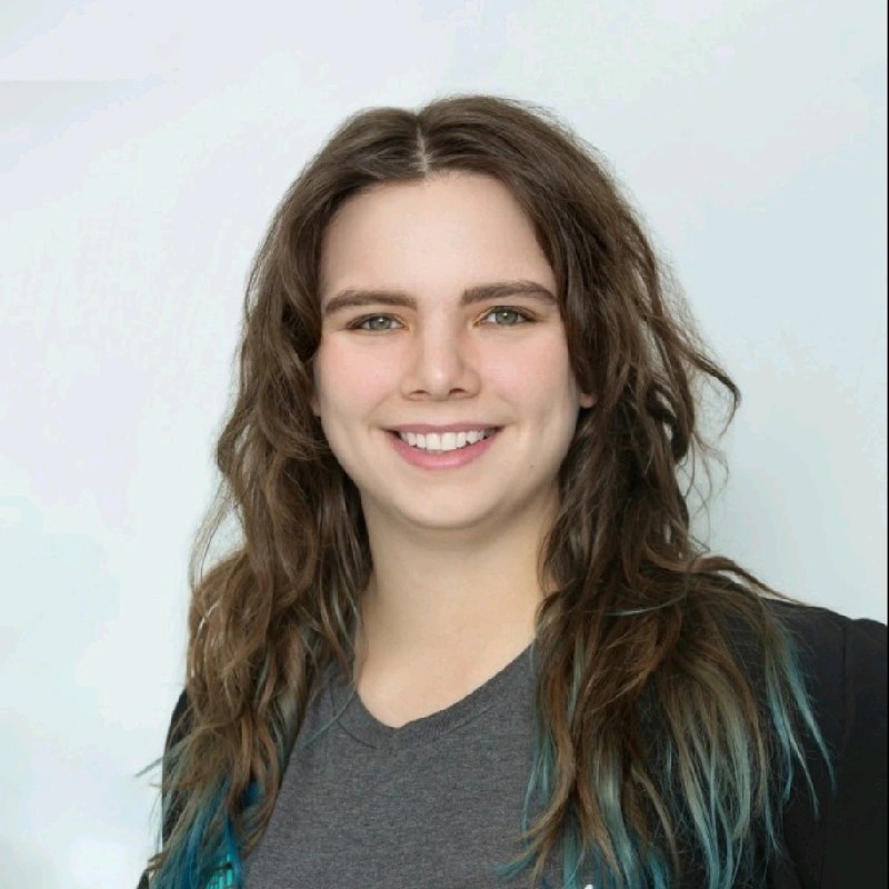 Professional headshot of a smiling person with long brown hair, wearing a gray shirt and dark blazer against a light background.